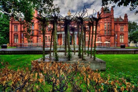 Sculpture. Five rows of slender tree trunks are set into a slab of concrete so their roots thrash against the sky. The concrete slab sits on a grassy lawn. A stately red brick building in the background nearly spans the width of the photograph.