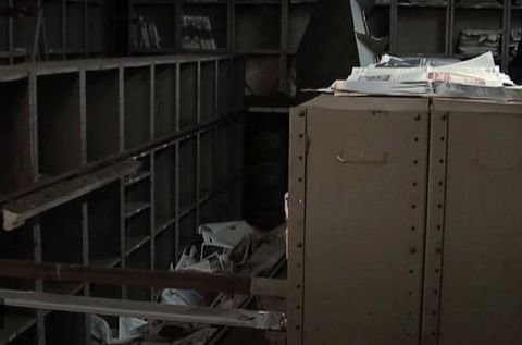Video still. Piles of loose sheets of newspaper are between and on top of metal shelves.