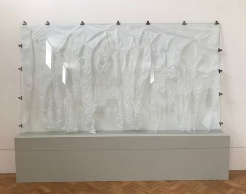 Installation. A tattered white cloth is displayed behind glass against a white wall. Fifteen bulldog clips hold the cloth to the glass, which is on a grey platform.